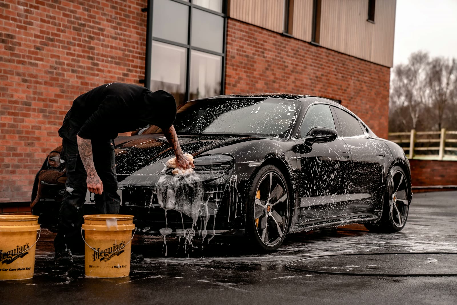 Detailer hand-washing a black Porsche covered in soap suds beside yellow buckets